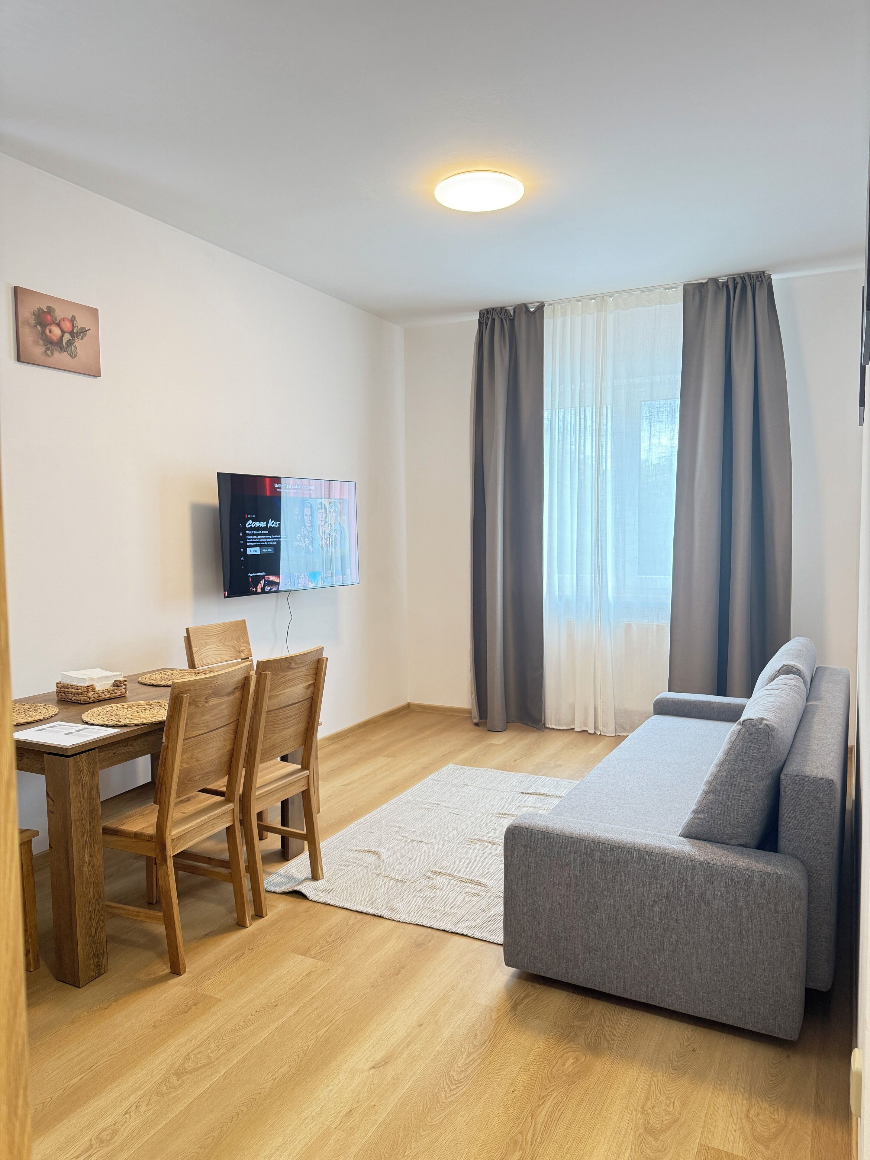 Ein Wohnzimmer mit einem Holzesstisch, Stühlen, einem grauen Sofa, einem an der Wand befestigten Flachbildfernseher und einem Fenster mit Vorhängen.
