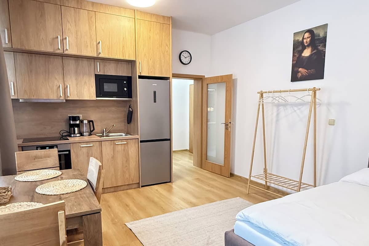 Une cuisine compacte avec des meubles en bois, un four à micro-ondes et un réfrigérateur. On y trouve une table à manger avec des chaises et un lit recouvert d’une couverture bleue.