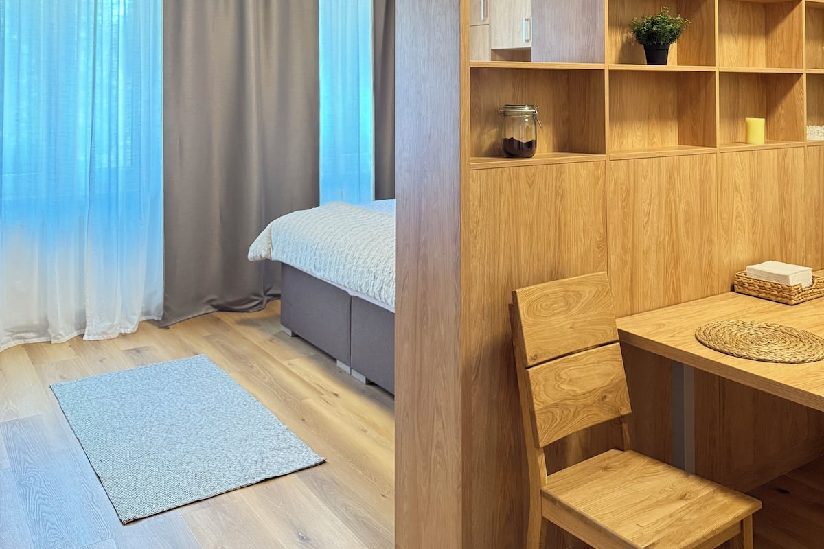 Une chambre à coucher moderne avec un lit, un bureau et une chaise. On y trouve une fenêtre avec des rideaux, une petite table avec une plante et une étagère avec des boîtes de rangement.