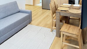 A modern kitchen with wooden cabinets, a stainless steel refrigerator, and a microwave. There is a dining table with chairs and a small wooden stool. A gray sofa and a white rug are in the living area. - Parkside Apartments Prague (Prague)