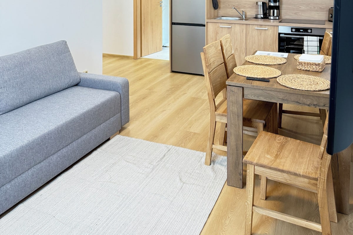 Une cuisine moderne avec des armoires en bois, un réfrigérateur en acier inoxydable et un four à micro-ondes. On y trouve une table à manger avec des chaises et un petit tabouret en bois. Un canapé gris et un tapis blanc se trouvent dans l’espace salon.