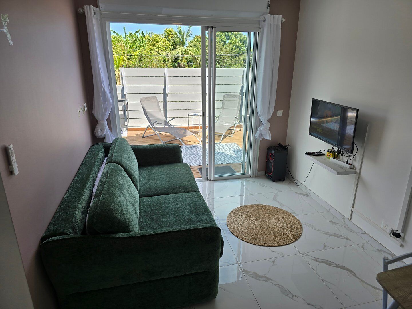 Un salon avec un canapé vert, un téléviseur et une porte-fenêtre coulissante donnant sur un espace extérieur comprenant une piscine et des chaises longues.