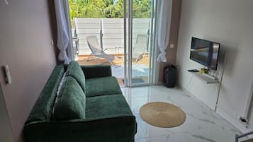 Un salon avec un canapé vert, un téléviseur et une porte-fenêtre coulissante donnant sur un espace extérieur comprenant une piscine et des chaises longues.