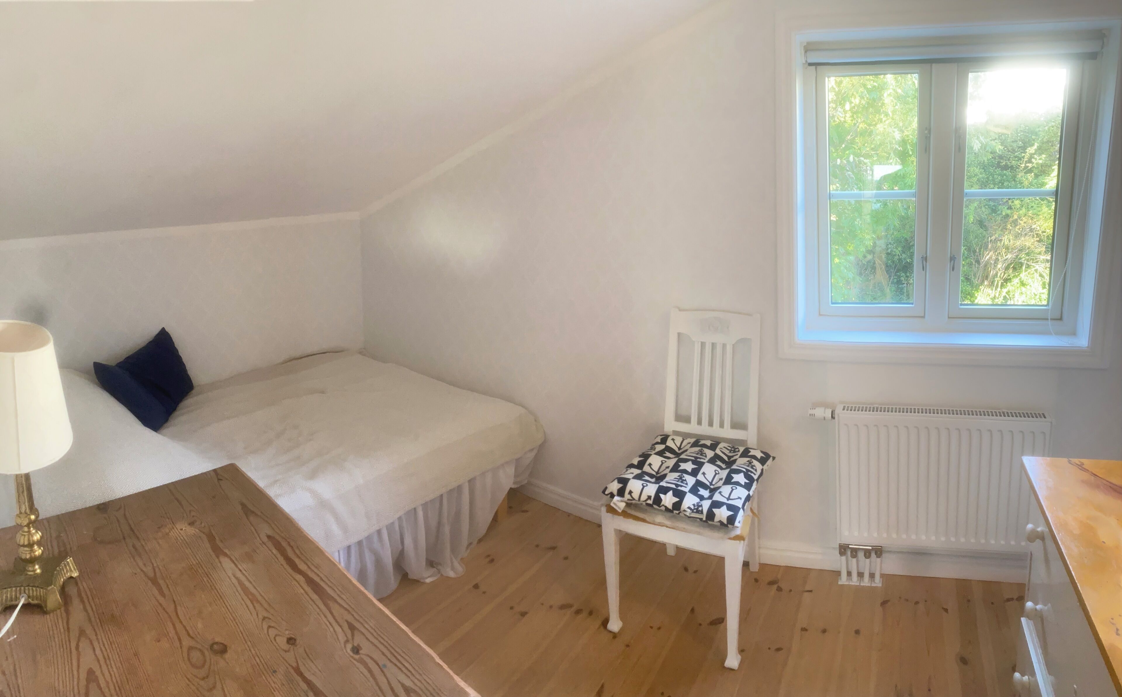 A bedroom with a single bed, a chair, a wooden dresser, and a window with a view of greenery.