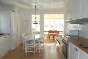 Eine Küche mit weißen Schränken, einer Holzausziehtischplatte, einem Esstisch mit Stühlen und Blick ins Freie durch große Fenster.