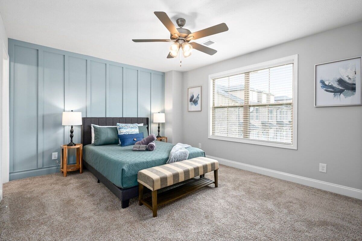 A bedroom with a bed, a ceiling fan, a window with blinds, and a wall with a painting.