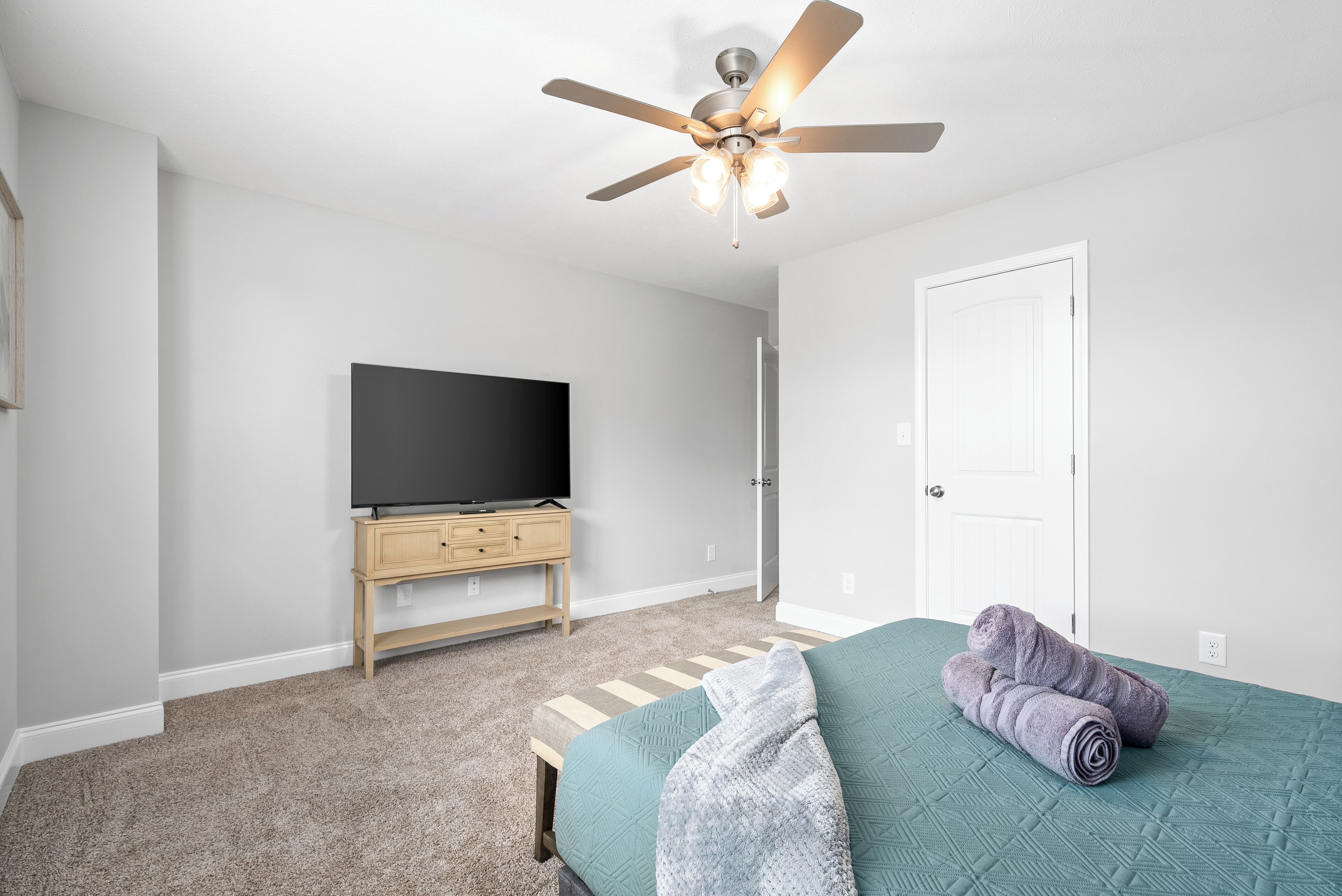 Um quarto com cama, ventilador de teto, TV sobre um suporte de madeira e uma porta.