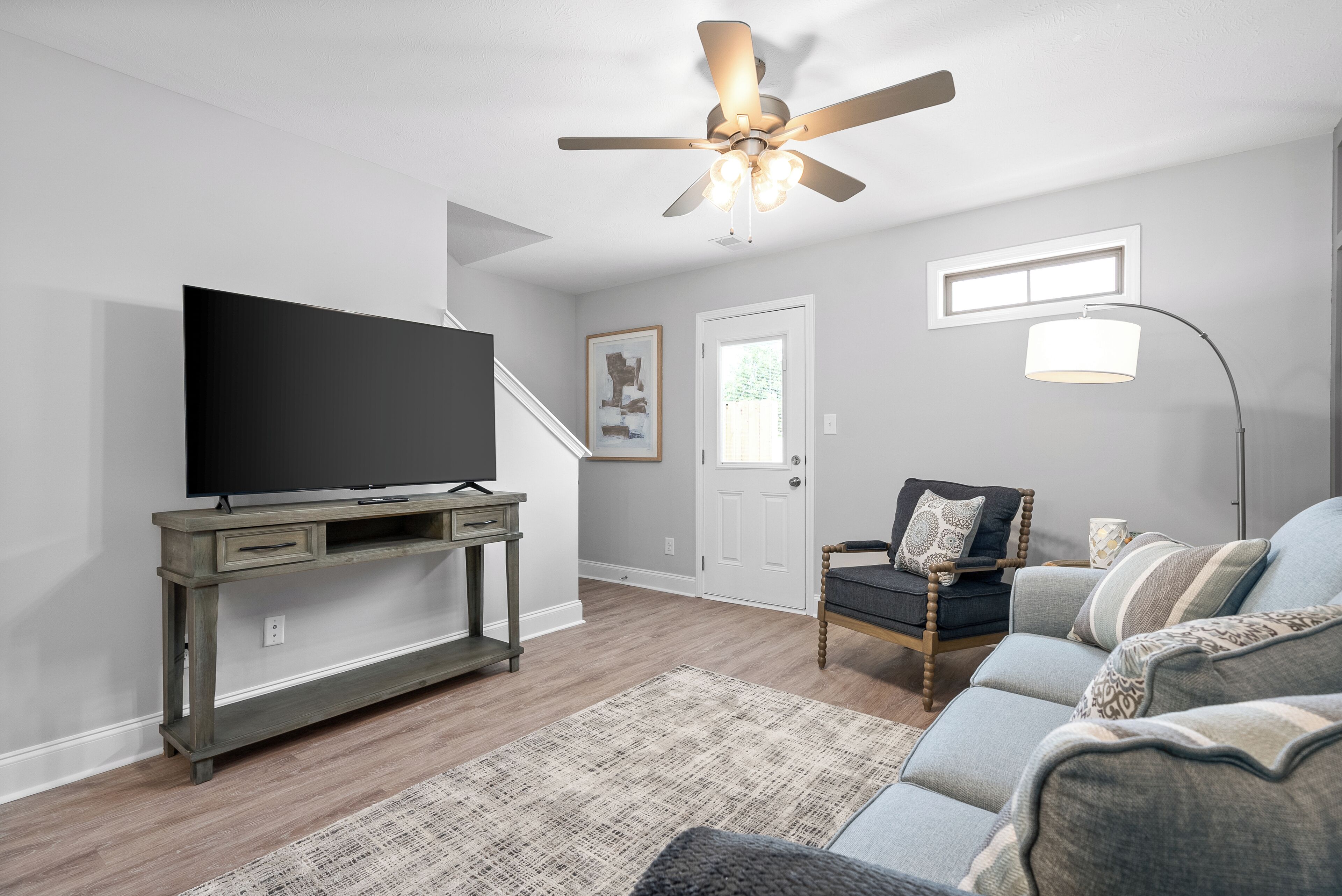 A living room with a flat-screen TV on a wooden stand, a ceiling fan, a sofa, a coffee table, and a rug.