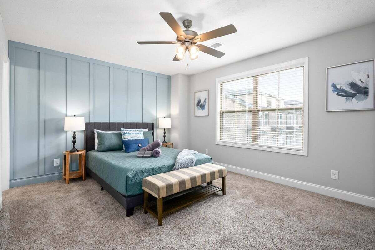 Un dormitorio con una cama, un ventilador de techo, una ventana con persianas y una pared con un cuadro.