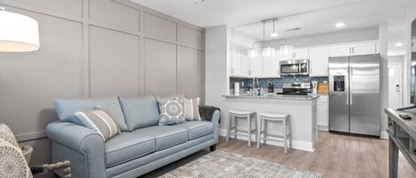 A modern living room with a grey sofa, patterned rug, and a kitchen area with white cabinets and stainless steel appliances.