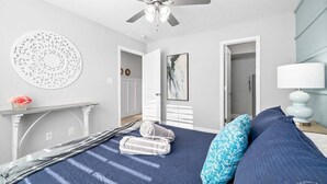Un dormitorio con cama, ventilador de techo, adorno circular en la pared, un cuadro, una lámpara y una mesita con un florero.