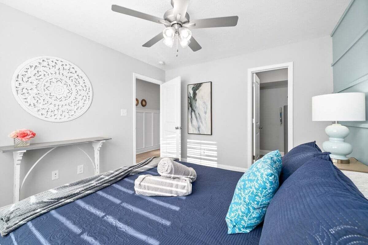 Un dormitorio con cama, ventilador de techo, adorno circular en la pared, un cuadro, una lámpara y una mesita con un florero.