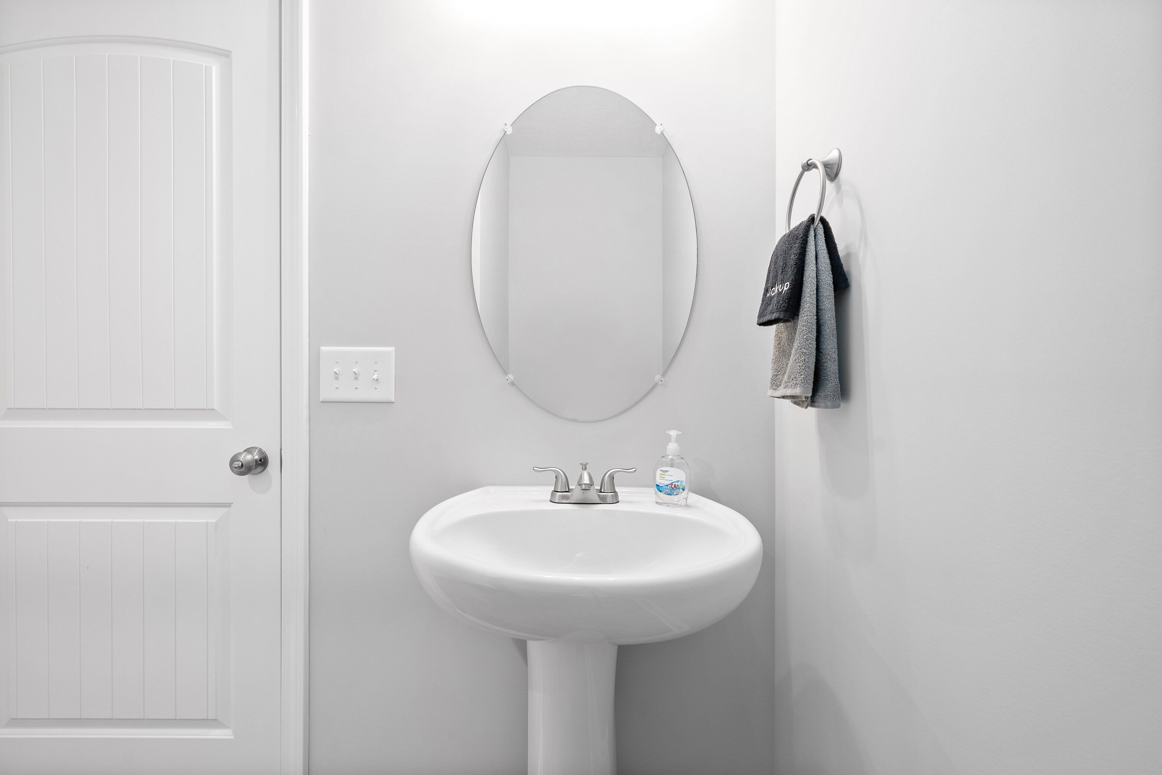 A white bathroom with a pedestal sink, a wall-mounted oval mirror, and a towel rack with a dark towel.