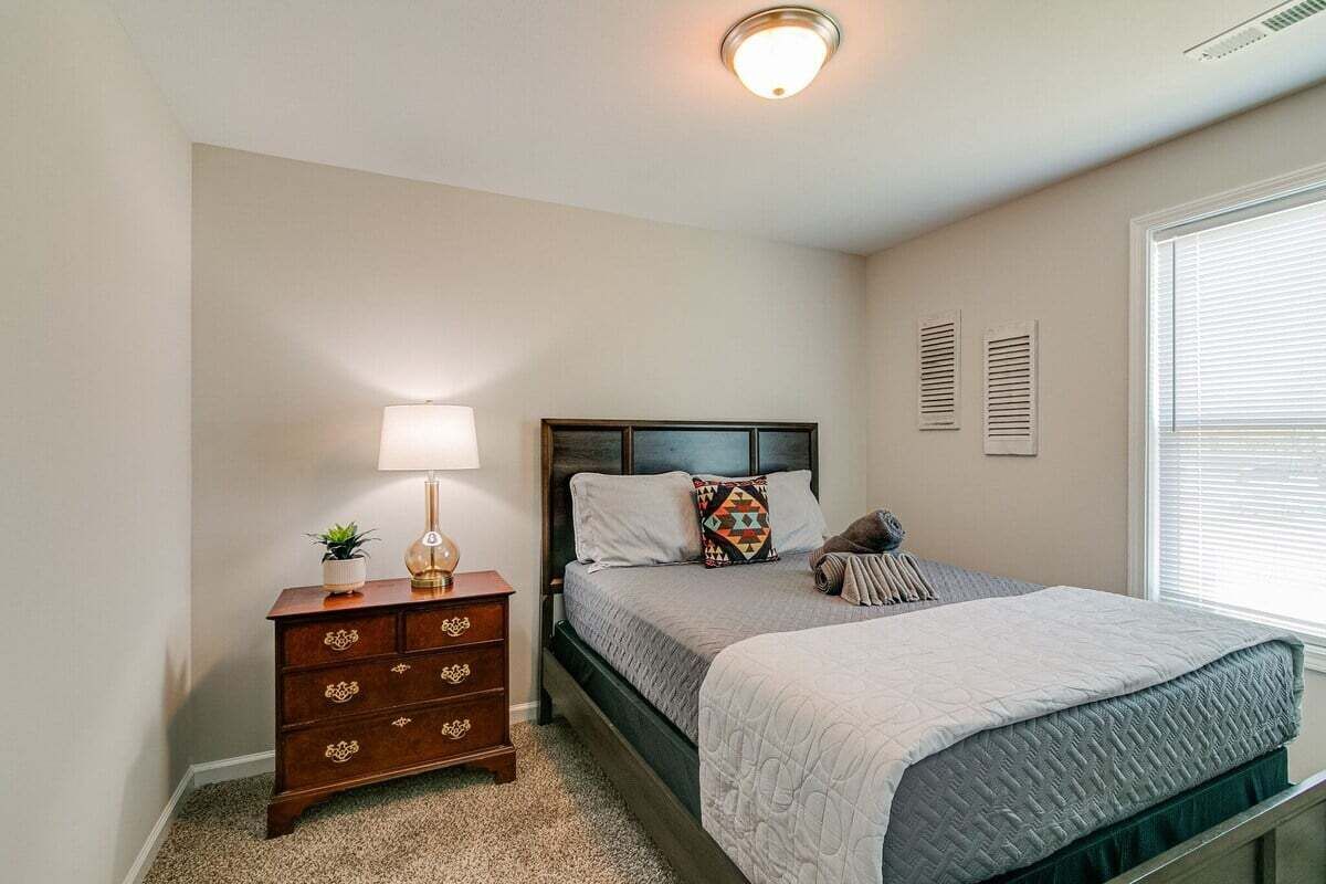 Quarto com cama, mesa de cabeceira com abajur e janela com persianas.