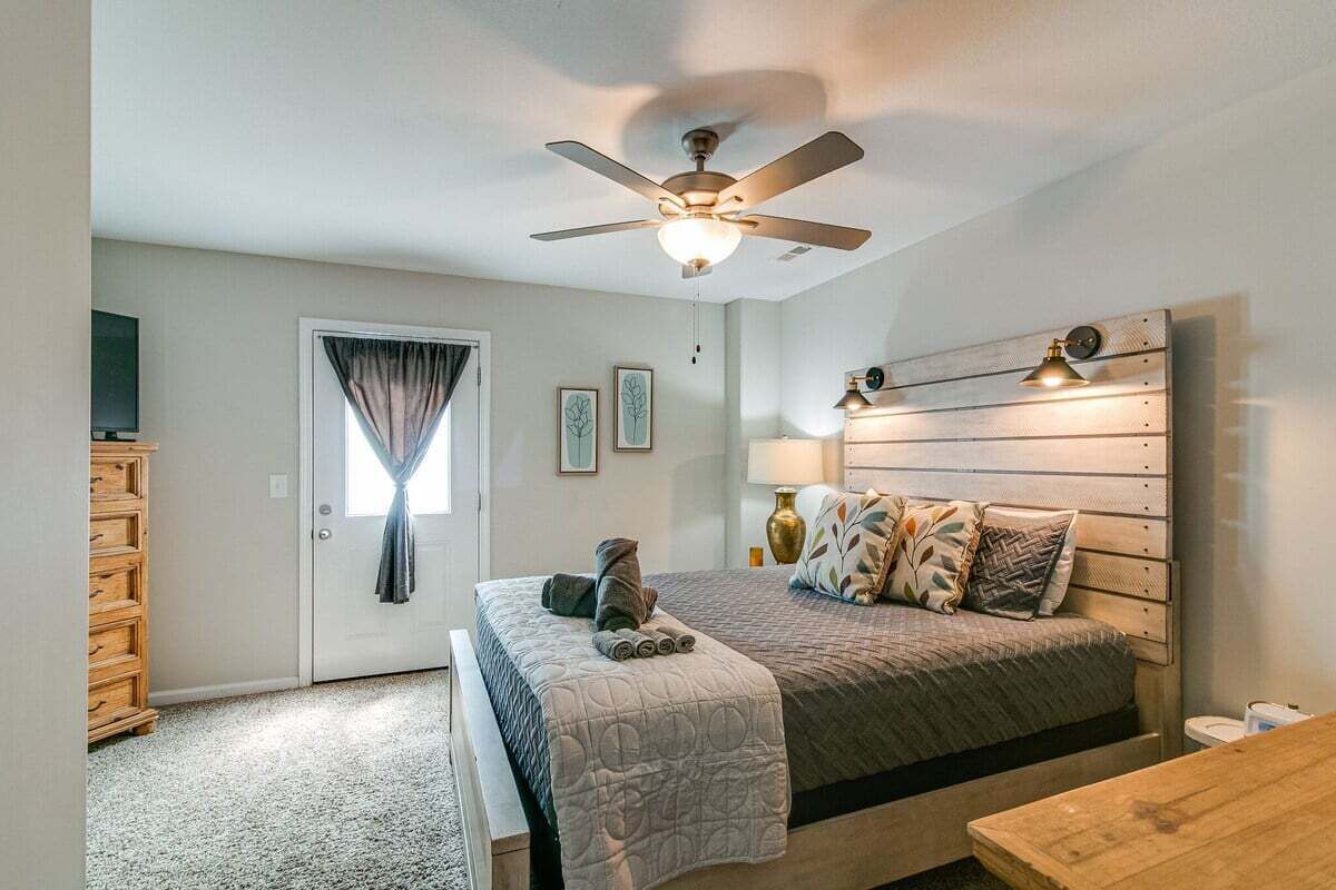 A bedroom with a bed, a ceiling fan, a bedside table, a lamp, and a window with curtains.