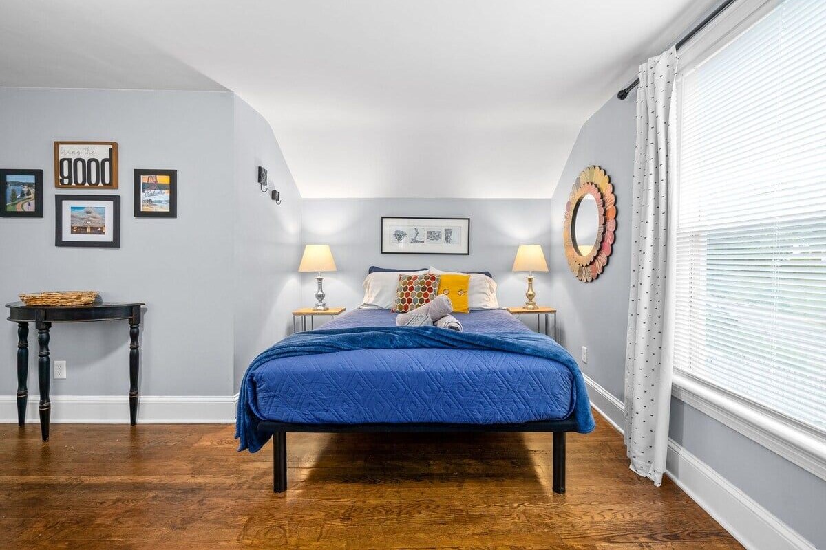 A bedroom with a bed, two bedside lamps, a small table, and a wall with framed pictures.