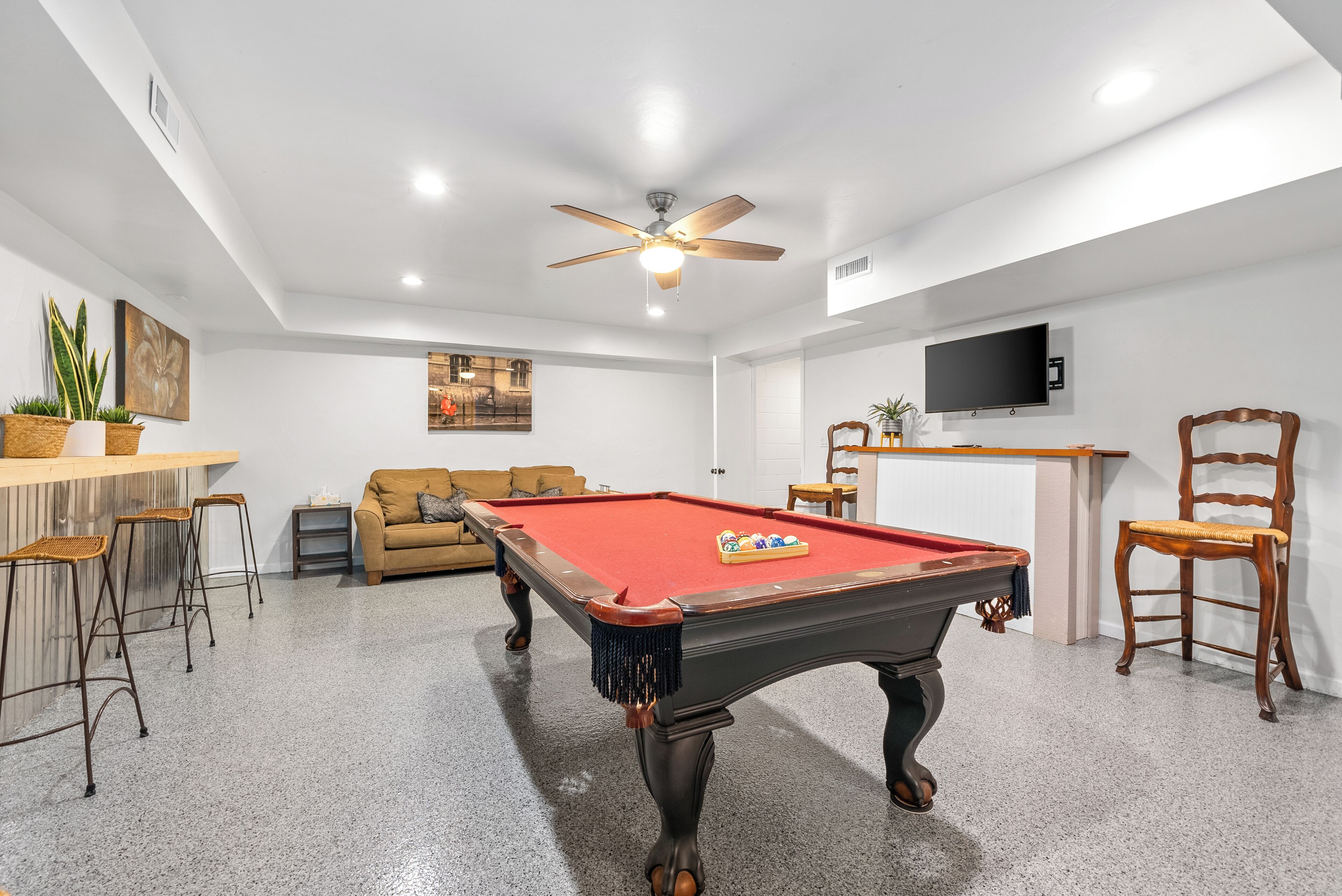 A pool table with a red felt surface, a ceiling fan, a sofa, and a television mounted on the wall.