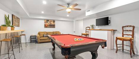 A pool table with a red felt surface, a ceiling fan, a sofa, and a television mounted on the wall.