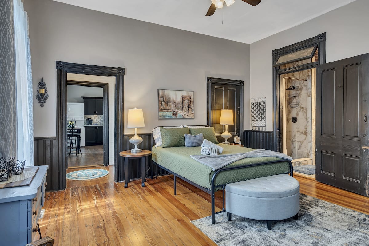 A bedroom with a bed, bedside lamps, a nightstand, a round ottoman, and a view into the kitchen.