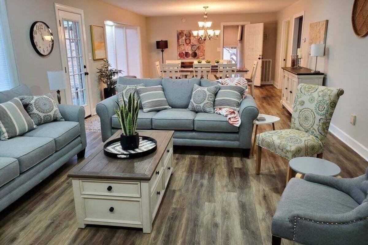 A living room with a grey sofa, a small side table, and a patterned armchair. There is a dining area in the background with a chandelier.