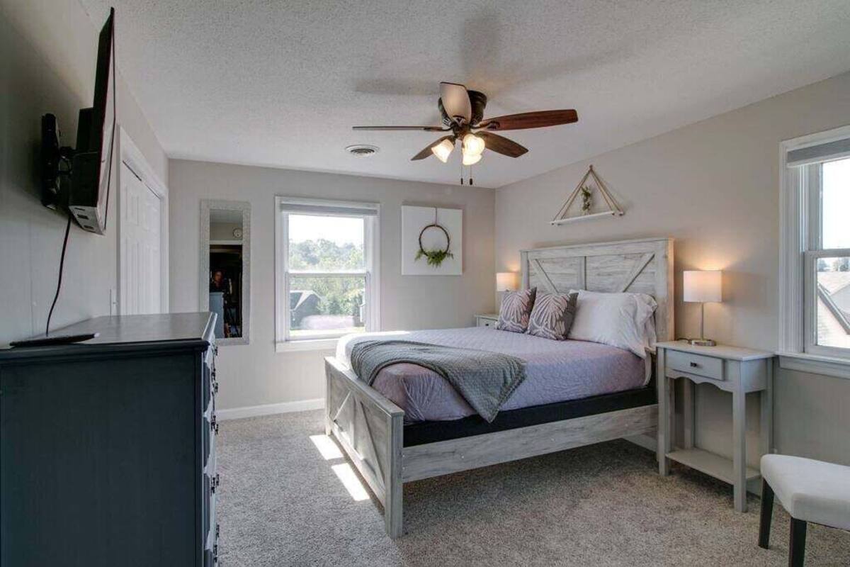 Une chambre à coucher comprenant un lit, des tables de chevet, un ventilateur de plafond, un téléviseur fixé au mur et une fenêtre.