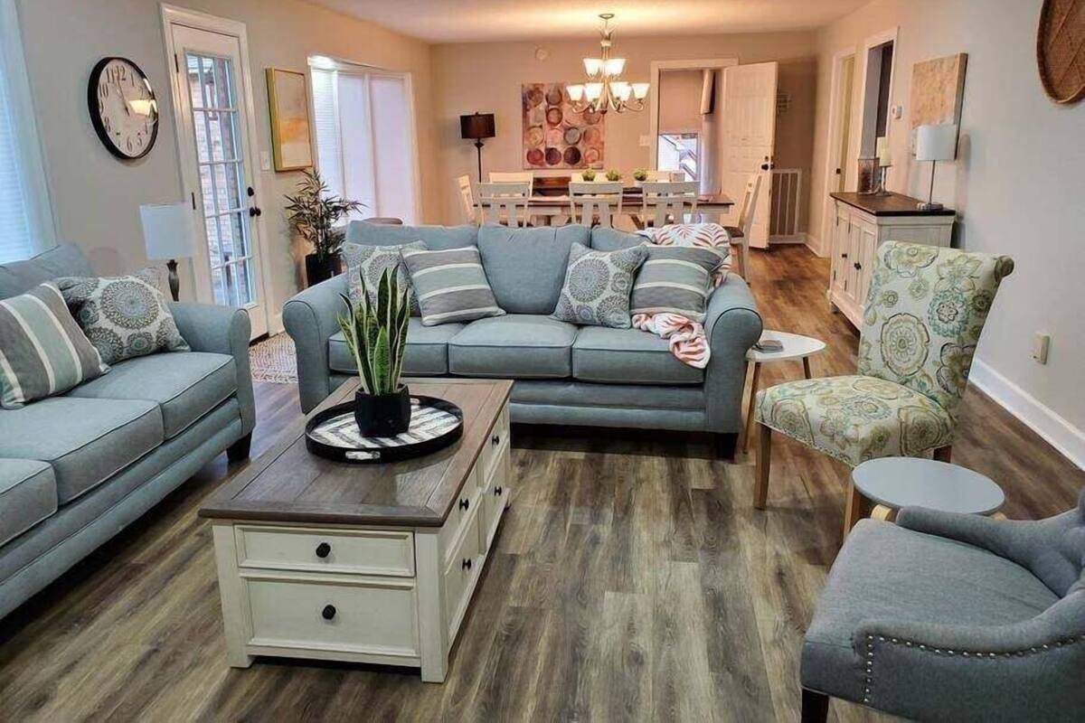 A living room with a grey sofa, a small side table, and a patterned armchair. There is a dining area in the background with a chandelier.