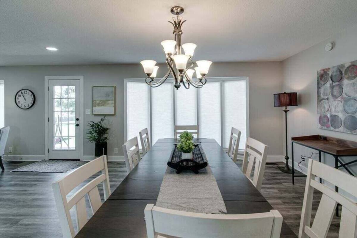 Une salle à manger avec une longue table, des chaises blanches, un lustre, une horloge et une porte avec des fenêtres.