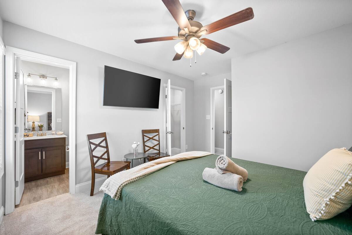 Une chambre avec une couvre-lit verte, un ventilateur de plafond, un téléviseur à écran plat, une coiffeuse avec un miroir et une chaise en bois.