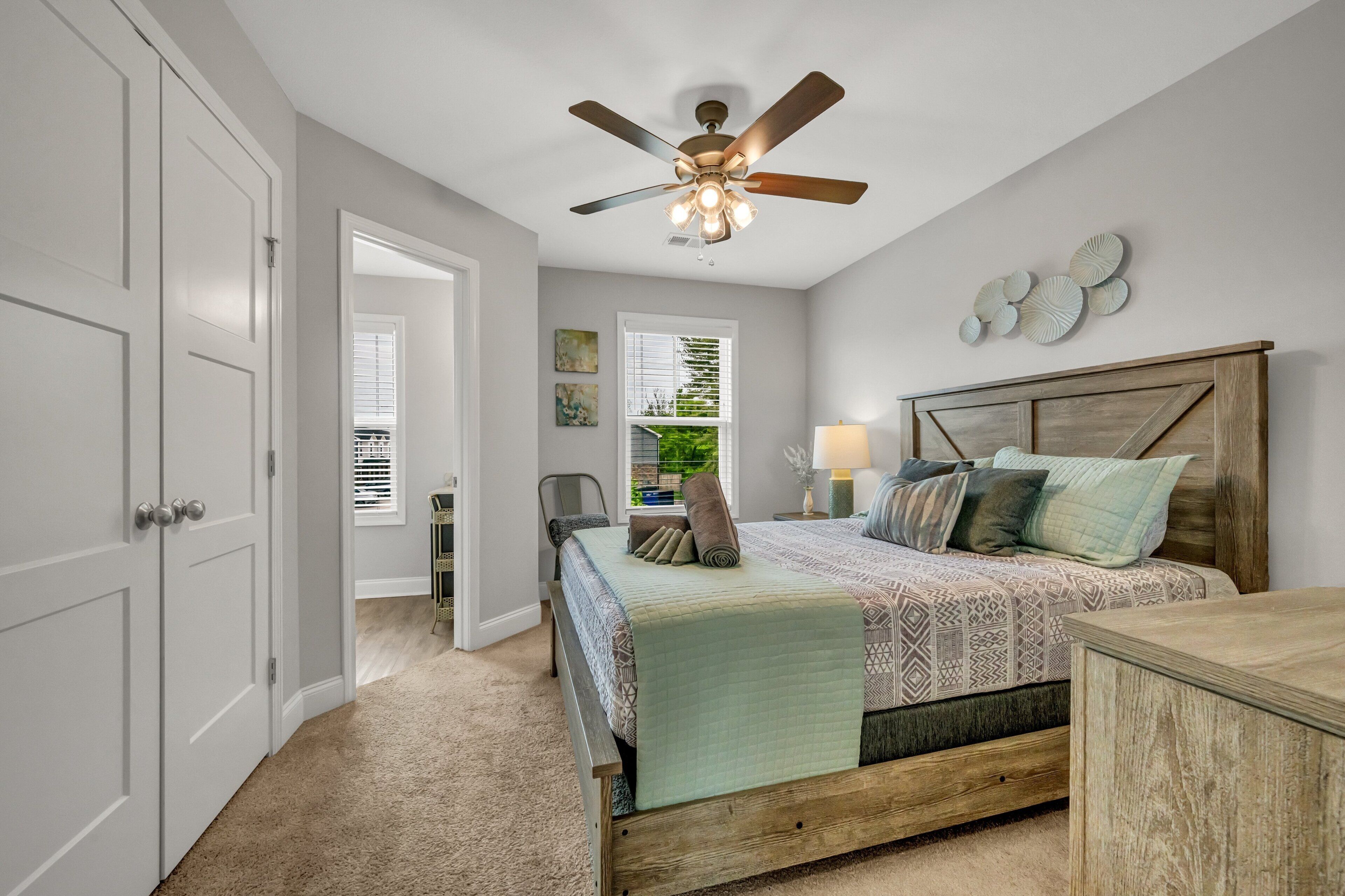 A bedroom with a bed, a ceiling fan, a nightstand, a lamp, and a view of the outdoors through a window.