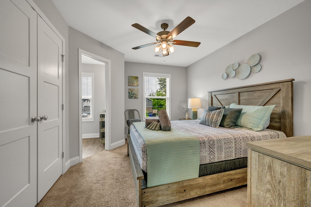 A bedroom with a bed, a ceiling fan, a nightstand, a lamp, and a view of the outdoors through a window.