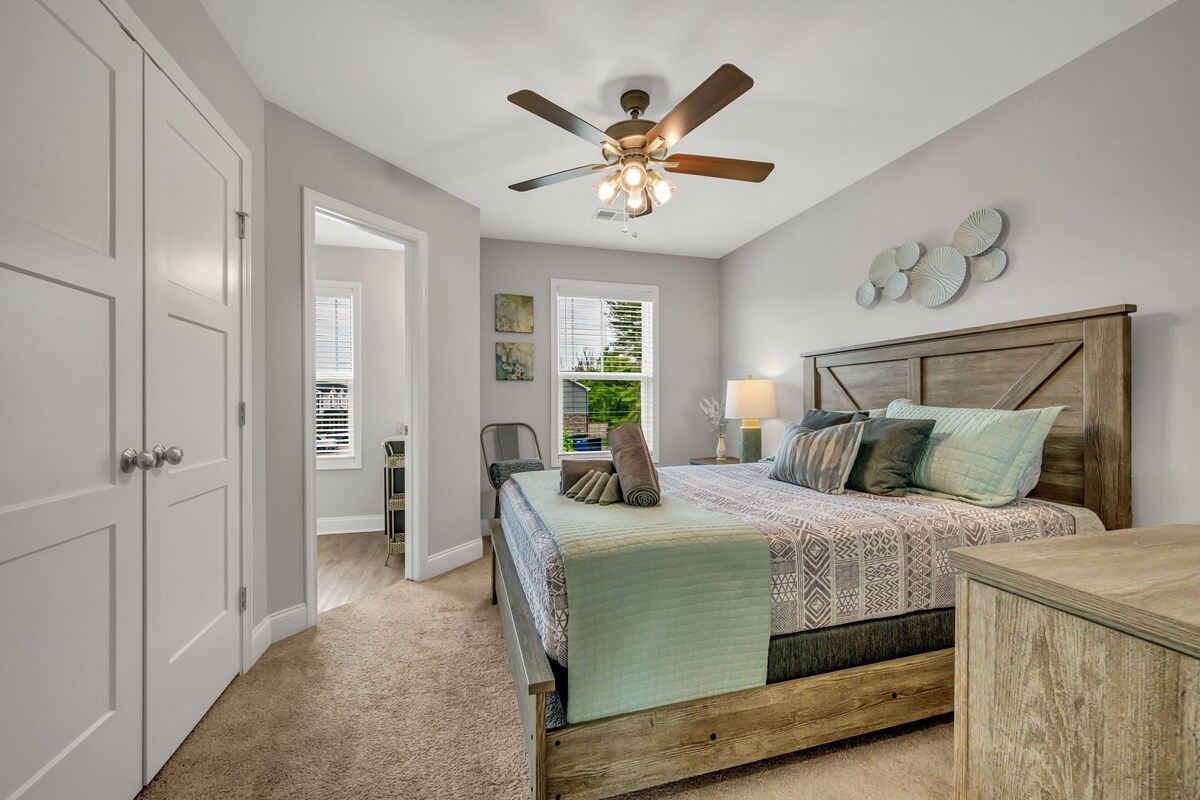 A bedroom with a bed, a ceiling fan, a nightstand, a lamp, and a view of the outdoors through a window.