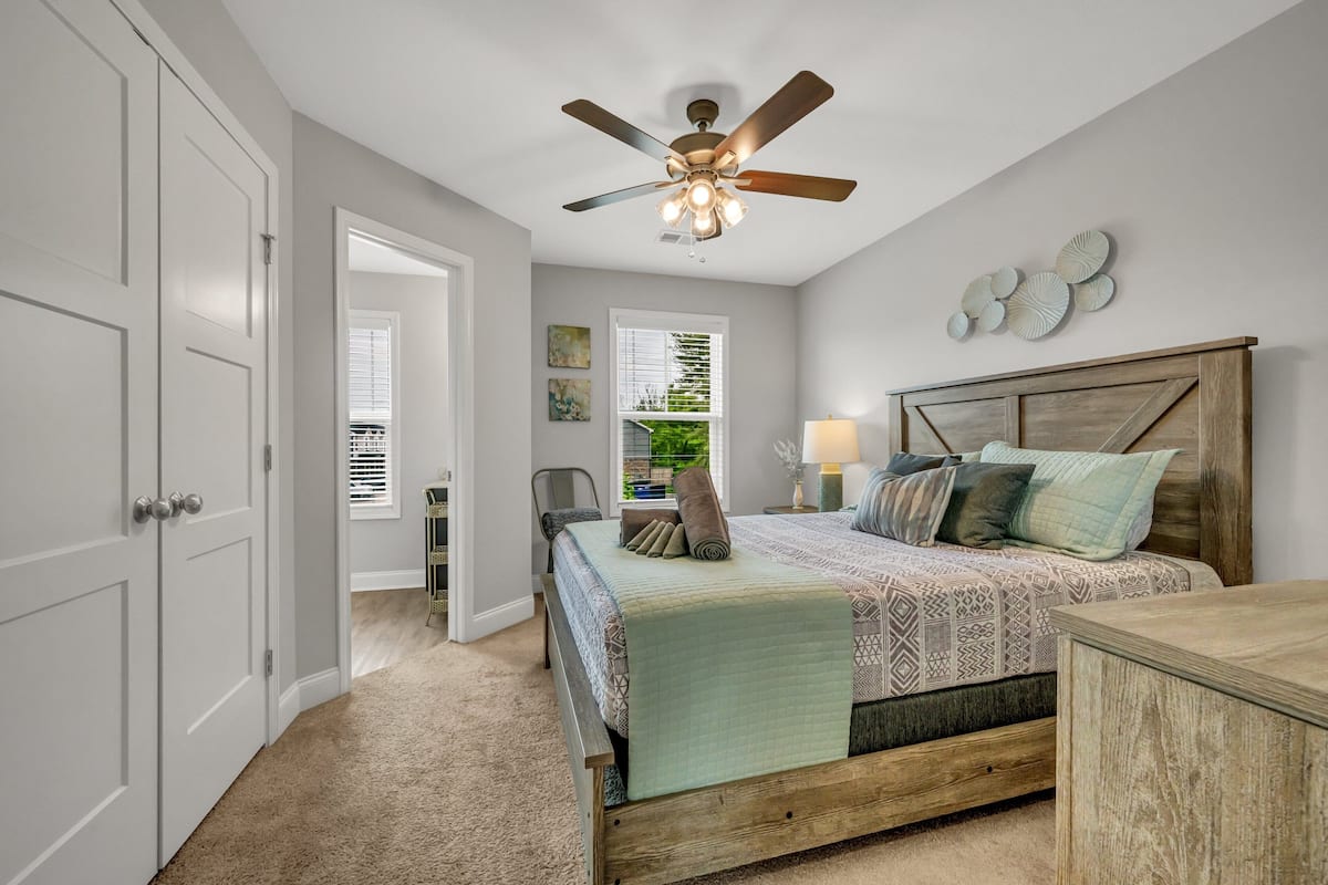 A bedroom with a bed, a ceiling fan, a nightstand, a lamp, and a view of the outdoors through a window.