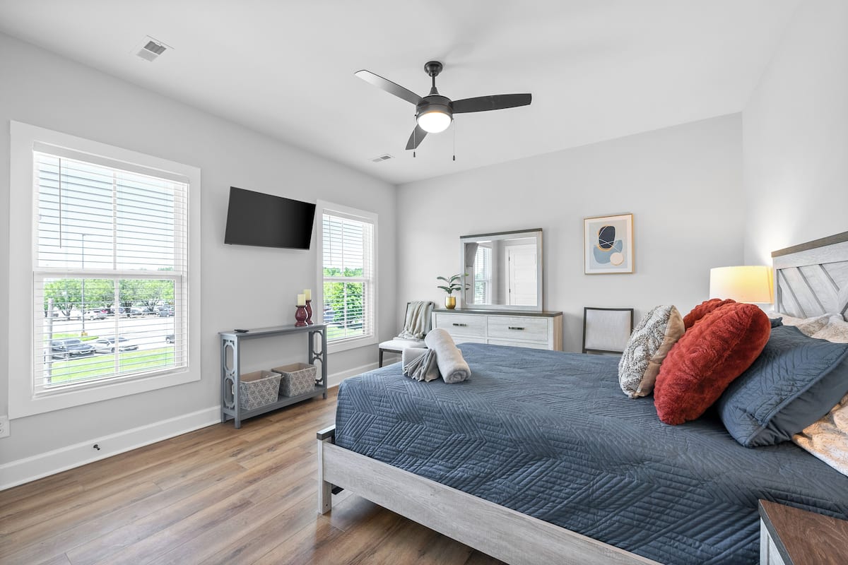 A bedroom with a bed, a ceiling fan, a TV, a dresser, a mirror, and a window with blinds.