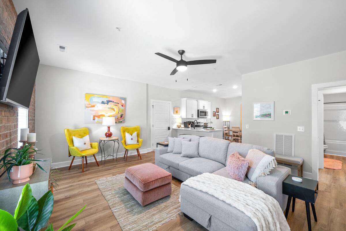 A modern living room with a grey sofa, ottoman, and yellow armchair. A flat-screen TV is mounted on the wall, and there is a ceiling fan.