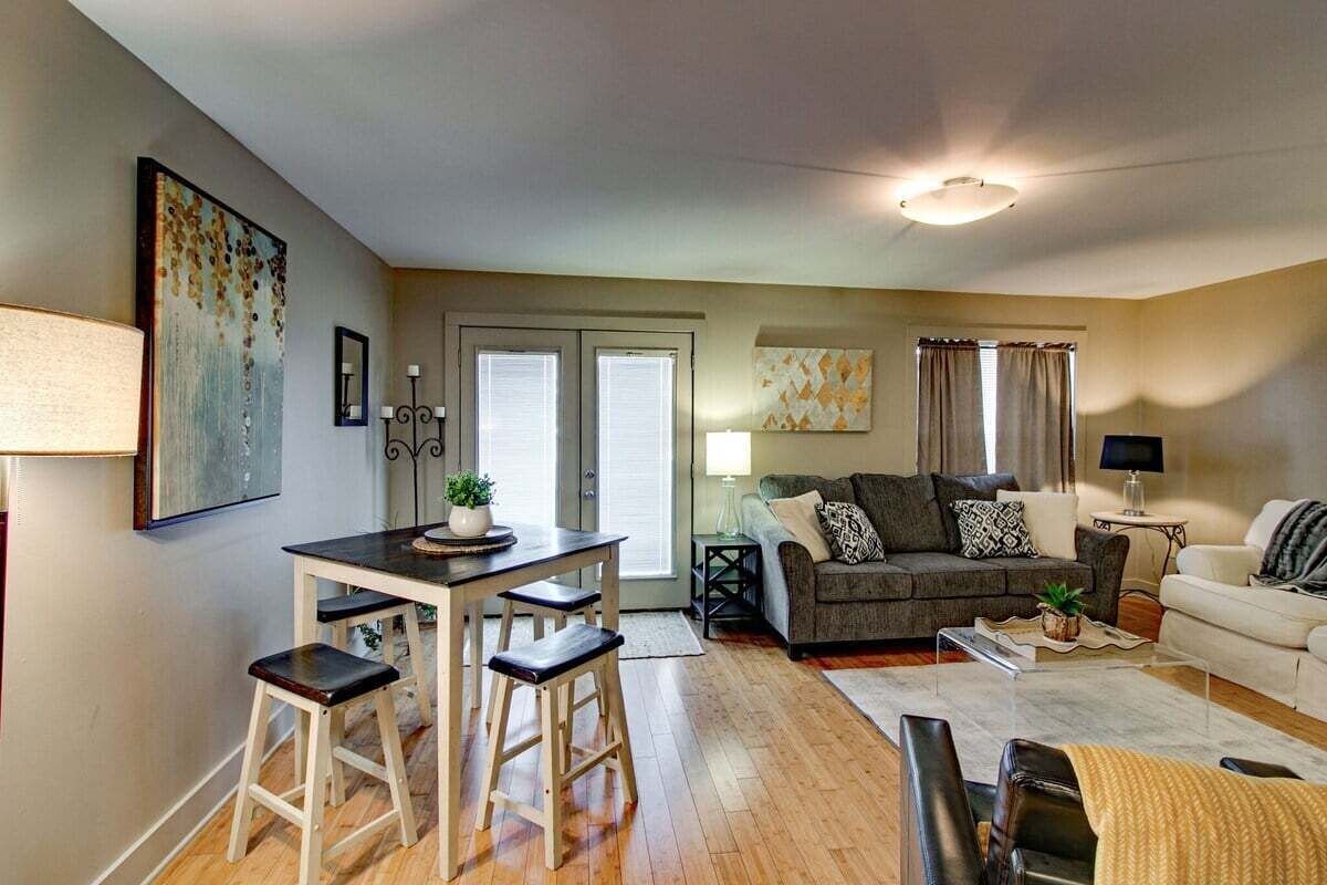 A living room with a sofa, a dining table with stools, and a coffee table. There is a window with curtains and a door with a glass panel.