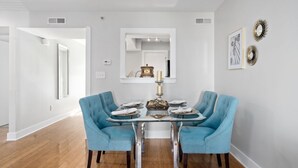 Un comedor con una mesa de vidrio, dos sillas tapizadas en azul, un centro de mesa decorativo y obras de arte colgadas en la pared.