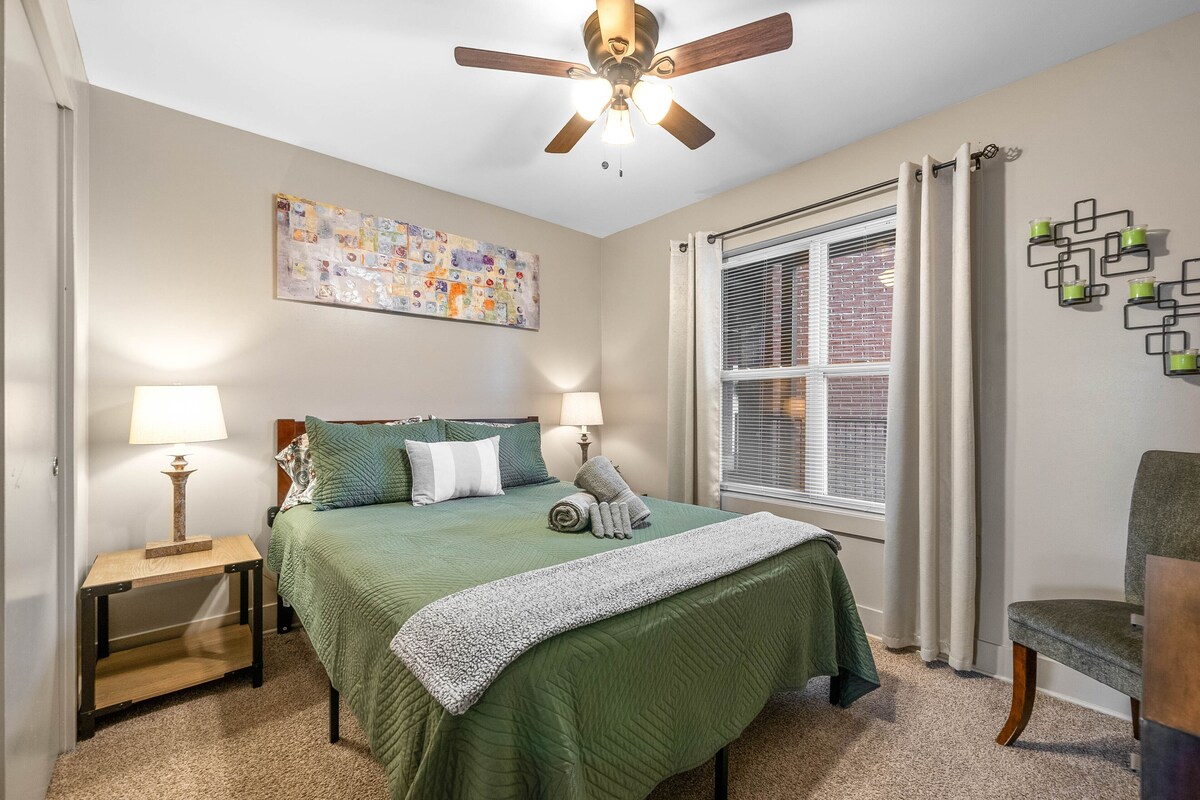 A bedroom with a bed, two bedside lamps, a ceiling fan, a window with blinds, a wall-mounted artwork, and a chair.