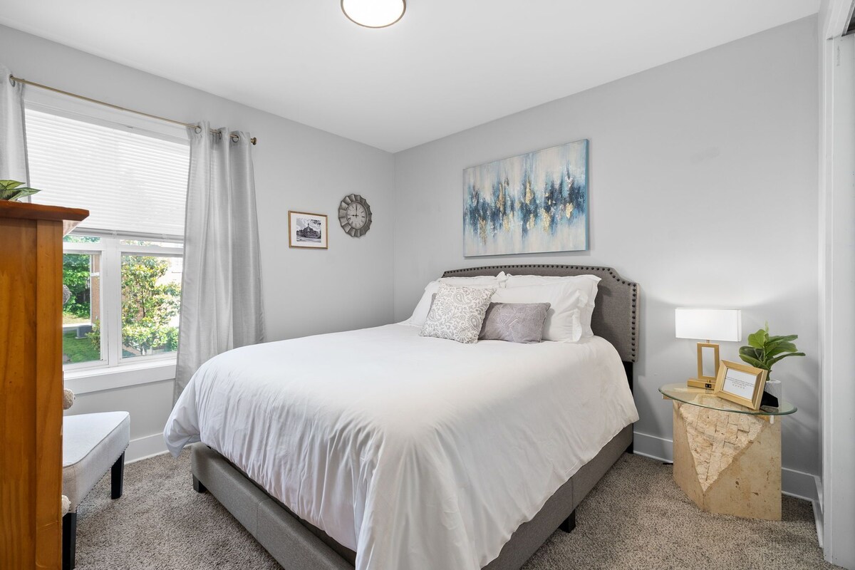 A bedroom with a large bed, a bedside table with a lamp, a window with curtains, and a painting on the wall.