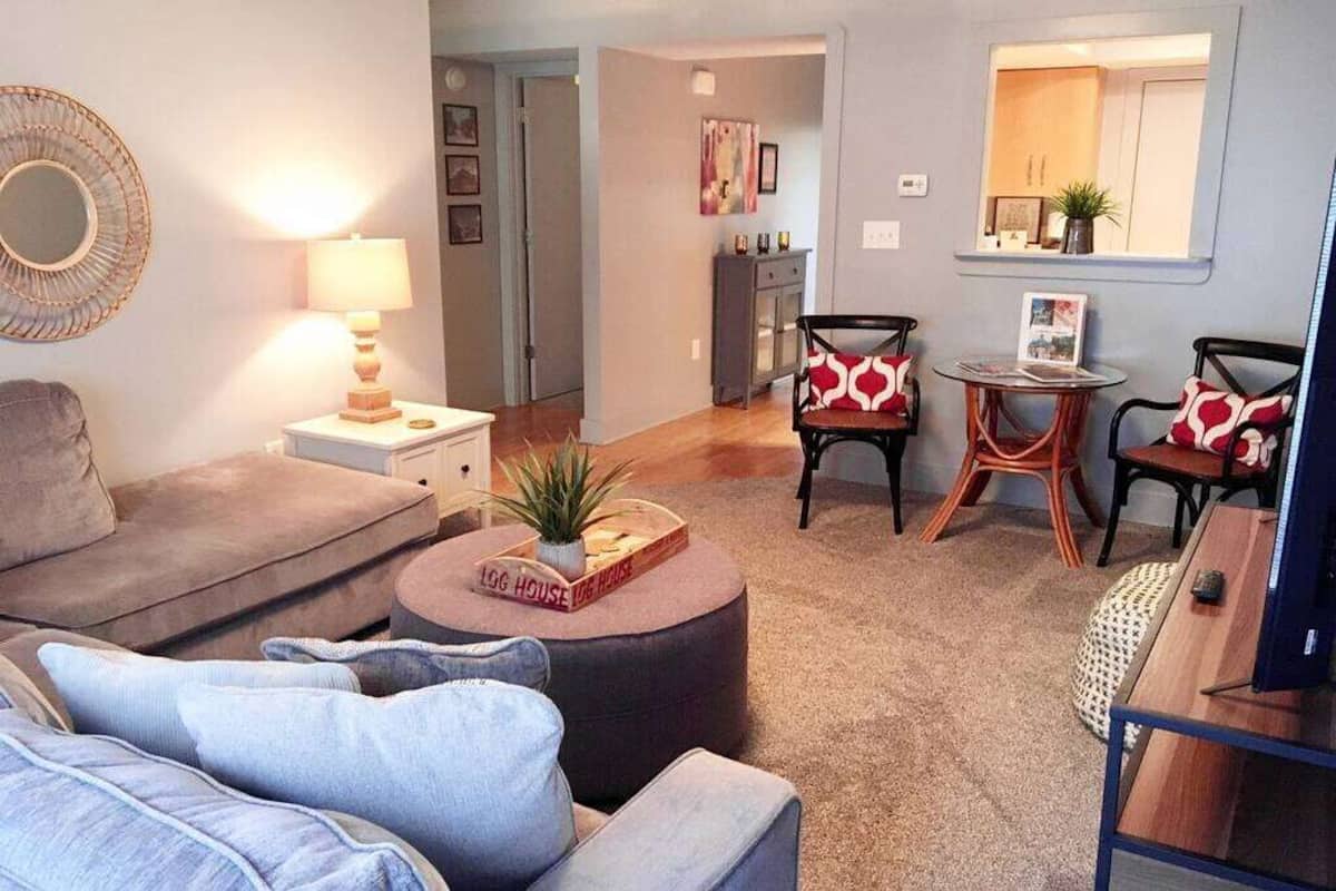 A living room with a sofa, a coffee table with a book and a plant, two chairs with patterned cushions, a small round table with a magazine, and a TV on a wooden stand.