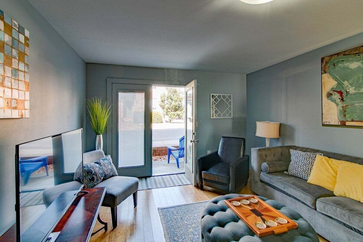 Un salon moderne avec un canapé, un fauteuil et une table basse. On y trouve une porte donnant sur l’extérieur, une plante en pot et une œuvre d’art accrochée au mur.