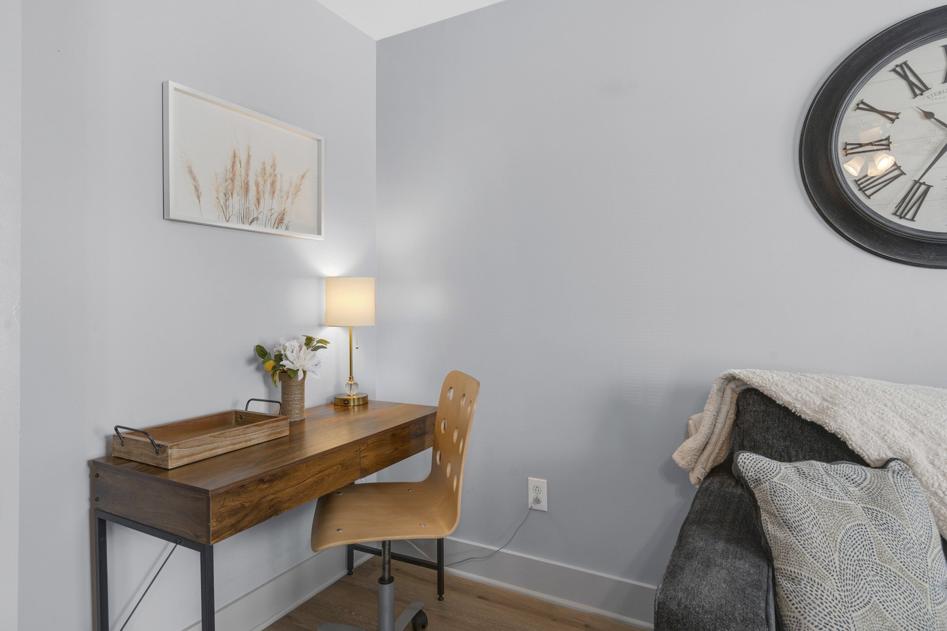 A room with a wooden desk, a chair, a lamp, a clock, and a couch with a blanket.