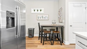 A modern kitchen with a stainless steel refrigerator, a wooden dining table with four chairs, a trash bin, and a window with potted plants. - Adam's Downtown Retreat w/ Rooftop Access (Clarksville)