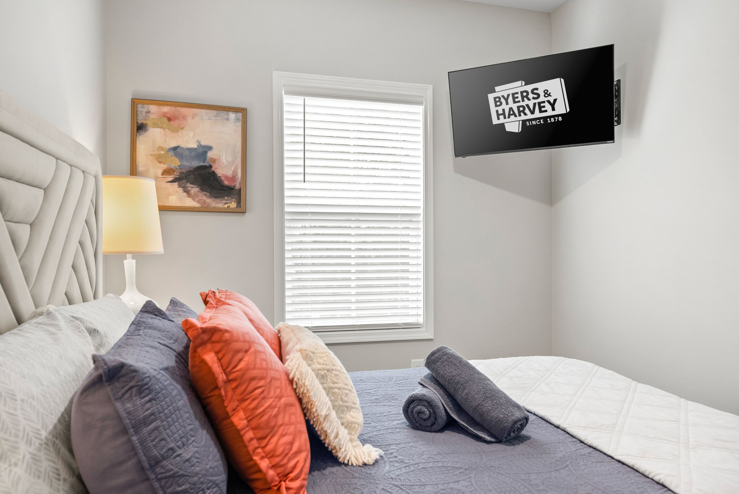 Une chambre à coucher avec un lit, des oreillers, une lampe, un tableau, une fenêtre avec des stores et un téléviseur fixé au mur.