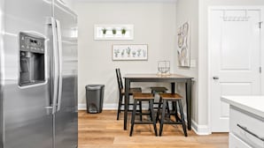 A modern kitchen with a stainless steel refrigerator, a dining table with four chairs, a trash bin, and a window with potted plants. - The Rooftop Haven of Downtown (Clarksville)