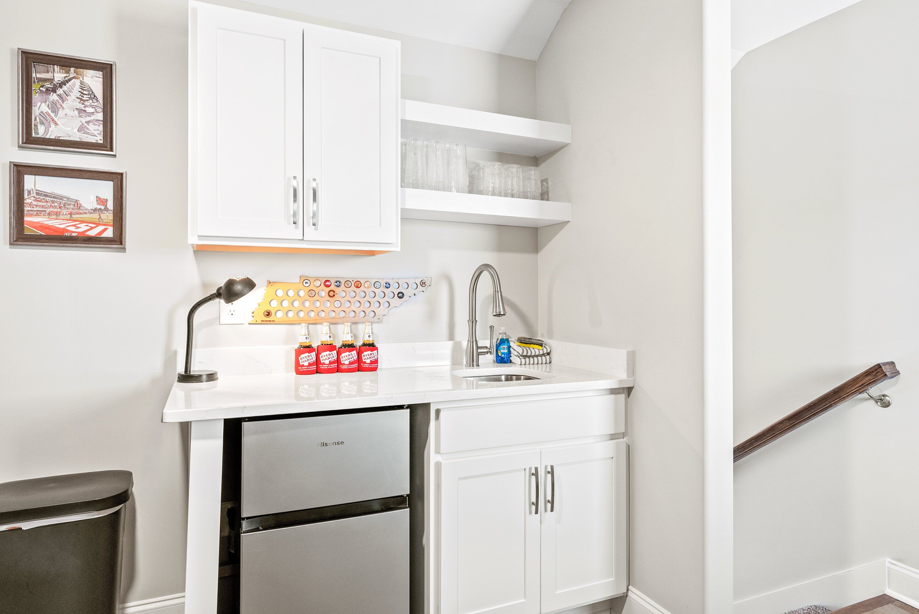 Una cocina con gabinetes blancos, un refrigerador de acero inoxidable, un lavamanos con grifo y una encimera con una cafetera y botellas.
