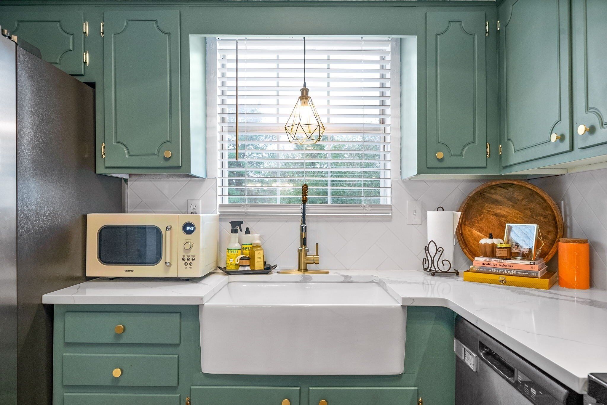 A kitchen with green cabinets, a white sink, a vintage microwave, and a window with blinds.