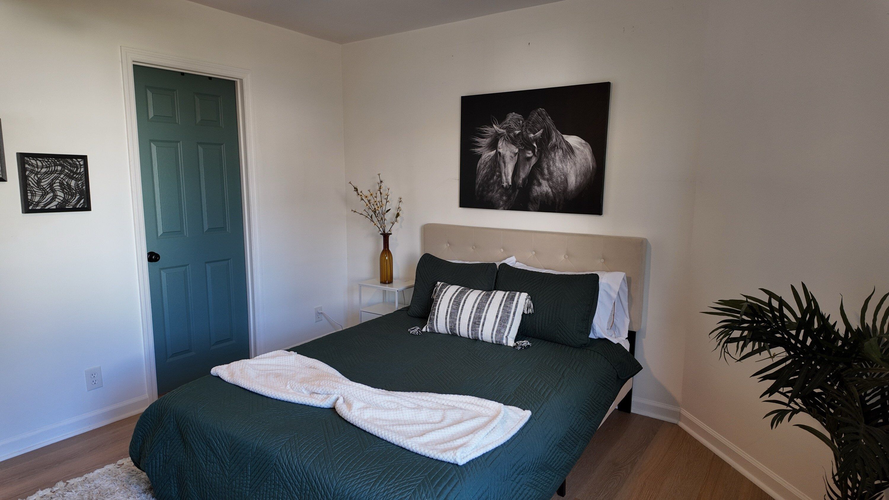 A bedroom with a bed, a door, a wall-mounted artwork, a vase with branches, and a potted plant.