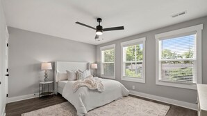 Une chambre à coucher avec un lit, des lampes de chevet, un ventilateur de plafond et de grandes fenêtres équipées de stores.