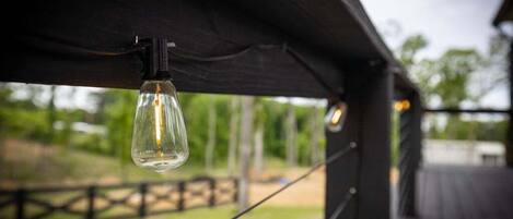 A hanging light bulb from a dark wooden structure with a view of a grassy area and trees in the background.