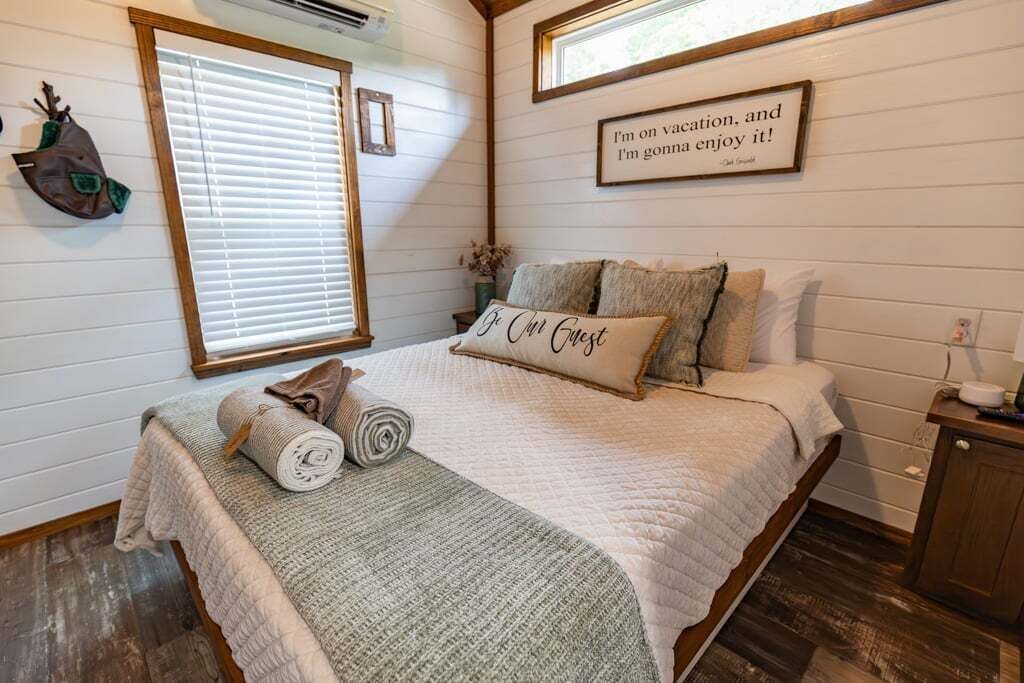 A cozy bedroom with a bed, pillows, a rolled blanket, a wall clock, and a quote sign.